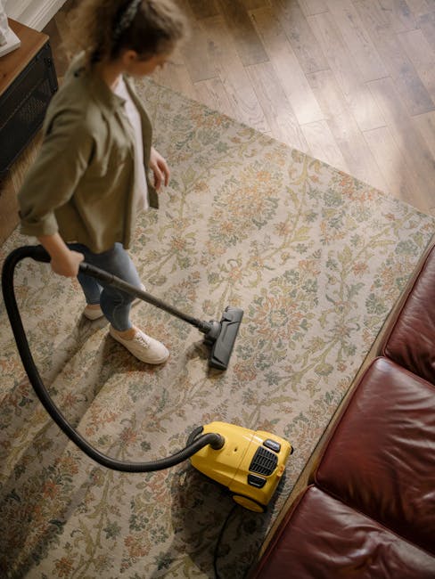 A woman is using a yellow and black vacuum cleaner to deep clean a patterned area rug in a living room. The room features wooden flooring adjacent to the rug and has a brown leather sofa positioned near the carpet. She is dressed in casual clothing, including a beige jacket and jeans. The lighting appears natural, and the surface of the rug looks fresh and dust-free, indicating professional sanitisation and carpet cleaning by Carpet Cleaning Kentish Town. The scene highlights domestic cleaning processes focused on surface care and hygiene.