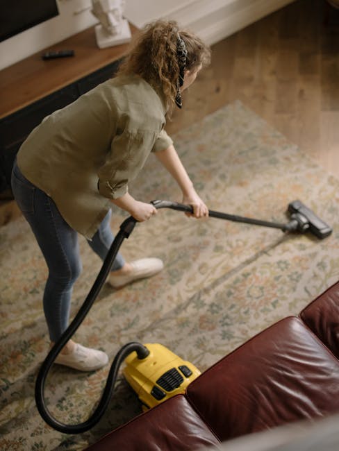 A professional cleaning specialist from Carpet Cleaning Kentish Town is performing deep carpet cleaning in a modern living room. The worker is dressed in full protective gear, including a white coverall, mask, and gloves, using a portable steam cleaning machine that emits steam or vapour onto the beige carpet. The room features a white wall, a wooden shelving unit with decorative items, and a large potted plant. Natural light illuminates the space, highlighting the freshly extracted moisture and foam from the carpet surface, indicating an intensive sanitisation process. Visible cleaning equipment and the pristine, wet carpet surface suggest thorough surface cleaning and deep cleaning aimed at removing dirt and stains, ensuring hygienic and spotless conditions. Carpet Cleaning Kentish Town specializes in professional domestic cleaning and surface sanitation, as demonstrated by this detailed carpet cleaning procedure.