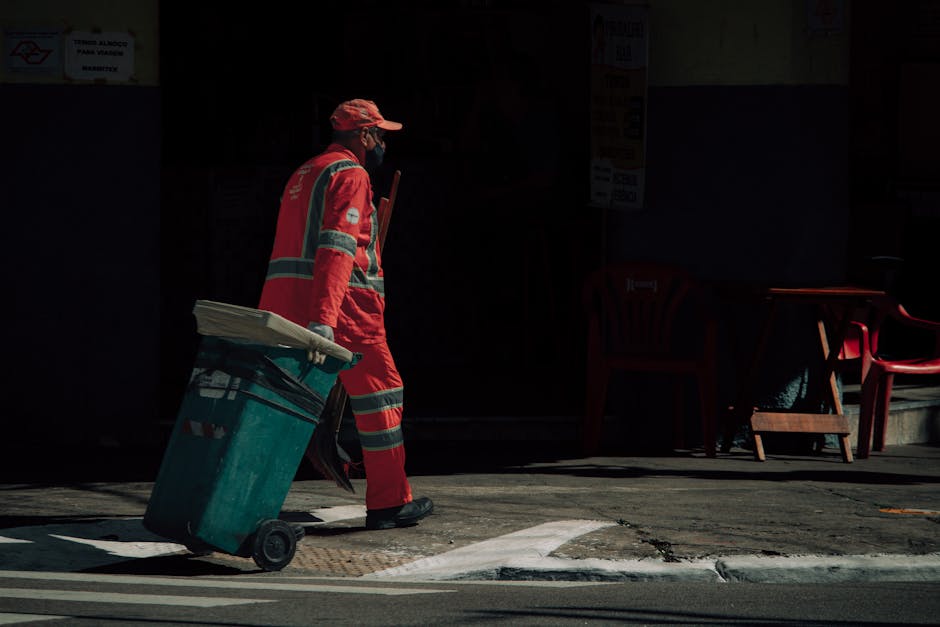 A person dressed in bright orange high-visibility workwear, including a jacket and trousers with reflective stripes, standing on a concrete sidewalk. They are facing a wall with a large pile of mixed 