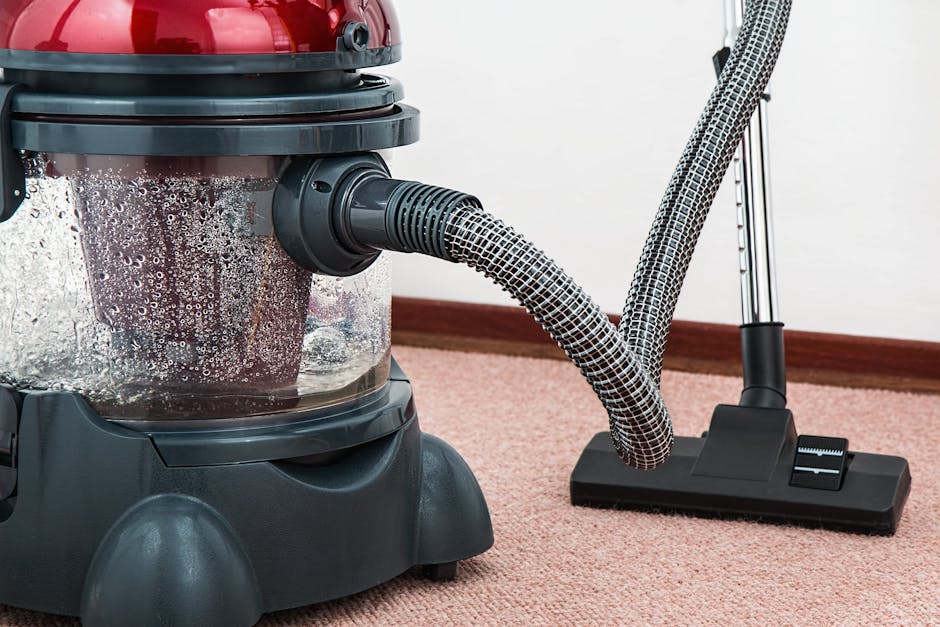 Close-up of a vacuum cleaner with a transparent dust canister filled with debris, positioned on a pink carpeted floor in a residential room. The vacuum has a flexible, patterned hose attached to the m