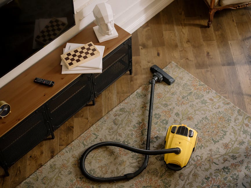 Close-up of a vacuum cleaner with a transparent dust canister filled with debris, positioned on a pink carpeted floor in a residential room. The vacuum has a flexible, patterned hose attached to the m