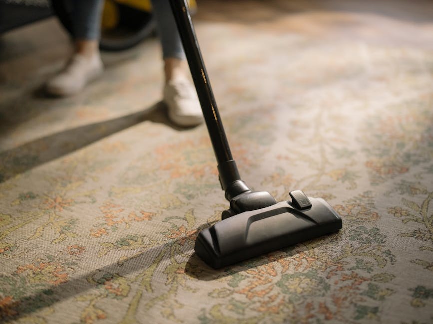 Close-up of a vacuum cleaner head with a black handle and motorized brush, actively cleaning a patterned carpet with floral motifs in beige, green, and orange tones. The vacuuming process appears thorough, with visible dust and debris being lifted from the carpet fibers. The room features soft, natural lighting that highlights the clean, well-maintained surface. In the background, a person wearing grey trousers and white sneakers is partially visible, standing on the carpet. This scene exemplifies professional carpet cleaning and surface maintenance, as provided by Carpet Cleaning Kentish Town, aligning with the focus on deep cleaning and hygiene for domestic and commercial spaces on the Fortess Road upholstery cleaning experts page.