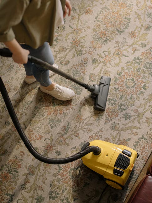 A person using a vacuum cleaner with a flexible hose attachment on a patterned, cream-colored carpet in a room with warm lighting. The carpet features intricate floral designs in shades of green, pink, and beige. The individual, wearing beige shoes, blue jeans, and a light-colored jacket, is performing deep cleaning or surface sanitisation as part of a domestic cleaning process. The yellow vacuum cleaner, of compact design, is positioned on the carpet nearby, connected to the hose. The scene emphasizes the thorough cleaning of the area by Carpet Cleaning Kentish Town, showcasing professional surface cleaning practices within a residential setting.