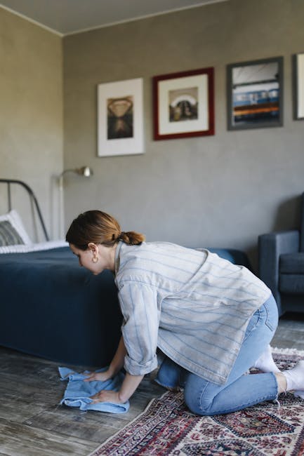 A woman with brown hair tied back, wearing a light blue striped shirt and blue jeans, is kneeling on a patterned rug and using a blue cloth to clean the wooden floor of a bedroom. The room features a dark blue bed with white bedding and a grey headboard, positioned against a beige wall decorated with three framed pictures. The lighting in the room is natural and soft, highlighting the clean and well-maintained surfaces. To the right, there is a blue upholstered armchair. The scene depicts domestic surface cleaning, emphasising hygiene and cleanliness, as part of the services offered by Carpet Cleaning Kentish Town, in preparation for end of tenancy or ongoing maintenance.