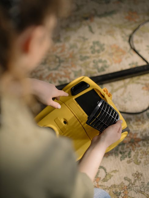 A person kneeling on a patterned carpet in a living room, using a yellow and black vacuum cleaner to perform surface cleaning. The individual is dressed casually in a beige jacket and blue jeans, and is holding the vacuum's handle while the machine's lid is open, revealing the filter and dust container inside. The room features wooden flooring in the background and soft ambient lighting, emphasizing the cleanliness and maintenance of the carpet. The scene highlights professional cleaning practices provided by Carpet Cleaning Kentish Town, matching the service focus of same day end of tenancy cleaning NW5 near Kentish Town station.