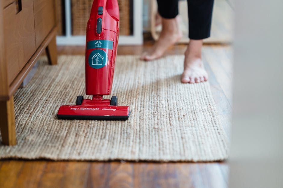A person using a red handheld vacuum cleaner to clean a beige textured carpet in a residential living room. The vacuum cleaner has a blue and white sticker indicating it is for surface cleaning and deep cleaning purposes. The individual, who is barefoot and wearing dark-colored pants, is standing on a wooden floor adjacent to the carpet, with part of their foot visible. The room features natural lighting, with a wooden sideboard or cabinet partially visible on the left. The carpet appears clean and well-maintained, highlighting the importance of hygiene and proper maintenance in domestic cleaning by Carpet Cleaning Kentish Town, especially when managing access issues related to cleaning services as discussed in the page titled 'What to know about access issues for Kentish Town cleaners.'